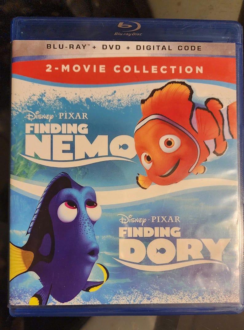 Puede incluir: Una caja de disco Blu-ray con las pel&iacute;culas de Disney Pixar Buscando a Nemo y Buscando a Dory. La caja tiene un fondo azul con im&aacute;genes de los personajes Nemo y Dory.