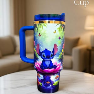 May include: A blue travel mug featuring Stitch from Lilo & Stitch, sitting on a pink lotus flower. The mug has a blue handle and lid, with a gold band. The background shows butterflies and tropical foliage. The text "Back of Cup" is visible.