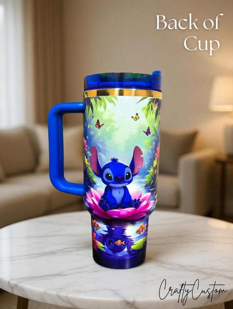 May include: A blue travel mug featuring Stitch from Lilo & Stitch, sitting on a pink lotus flower. The mug has a blue handle and lid, with a gold band. The background shows butterflies and tropical foliage. The text "Back of Cup" is visible.