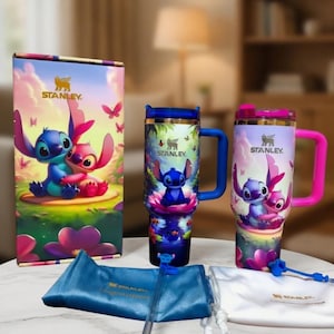 May include: A Stanley travel tumbler set featuring a colorful design of the characters Stitch and Angel. The set includes a tumbler with a blue handle, a tumbler with a pink handle, a box, a blue bag, and a white bag.