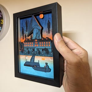 May include: A framed paper art piece with a layered design, showing a surreal scene with a building, figures, and a boat on water. The artwork has a dark frame, a blue and orange sky, and a dark pier. The overall theme is a dark, mysterious landscape.