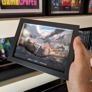 Può includere: Un'opera d'arte 3D incorniciata raffigurante una scena di guerra con carri armati, aerei e soldati. L'opera d'arte è in una cornice nera, con le parole "HELL LET LOOSE" in basso. Altre opere d'arte incorniciate con il testo "PixelArt GameCrafts" sono visibili sullo sfondo.