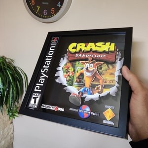 Puede incluir: Una carátula de videojuego enmarcada para el juego de PlayStation Crash Bandicoot. La carátula presenta un personaje de dibujos animados, Crash Bandicoot, corriendo por una escena de la jungla. El título del juego está escrito en letras amarillas con un contorno negro. La clasificación del juego es T para adolescentes. El juego es publicado por Sony Computer Entertainment y desarrollado por Naughty Dog.