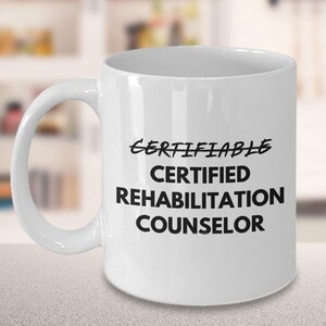 May include: White ceramic mug with the words "CERTIFIABLE CERTIFIED REHABILITATION COUNSELOR" printed in black. The mug has a curved handle and is set against a blurred background.