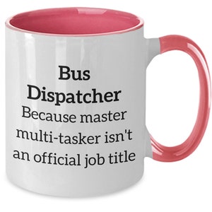 May include: White ceramic coffee mug with a pink rim and handle. The mug has black text that reads "Bus Dispatcher Because master multi-tasker isn't an official job title."