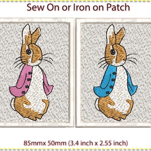 Op de afbeelding: Twee geborduurde strijk- of naai-patches met Peter Rabbit. Eén patch heeft Peter Rabbit met een roze jasje en de andere heeft Peter Rabbit met een blauwe jas. De patches zijn 85 mm x 50 mm.