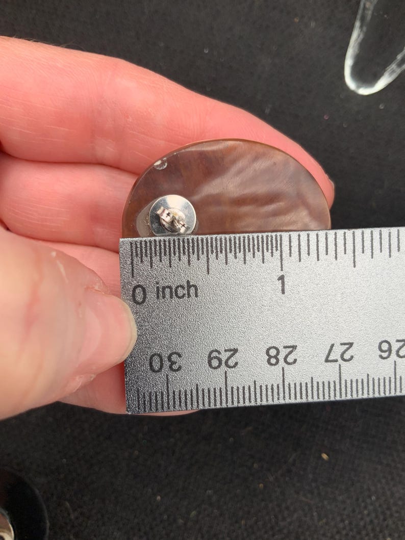 May include: A brown, semi-circular earring with a silver post, measuring approximately 1 inch (2.5 cm) in diameter. The earring is held up to a ruler for measurement, showing its size.