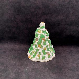 May include: A conical sculpture shaped like a tree, featuring a white textured base resembling frosting or snow, decorated with numerous small, translucent green and amber-colored glass or resin pieces embedded throughout the surface.