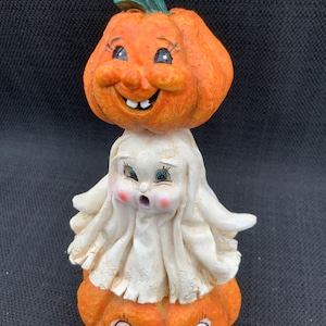 Puede incluir: Una caprichosa figura de Halloween con una cabeza de calabaza naranja sonriente con un tallo verde, encima de una figura de fantasma blanco. El fantasma está posado sobre una base de calabaza más pequeña, creando un diseño apilado. Detalles pintados y expresión alegre.