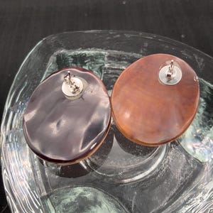 May include: Two oval-shaped stud earrings. One earring is a dark brown color, and the other is a lighter brown. Both earrings have a silver-colored post and backing. The earrings are displayed in a clear glass dish.