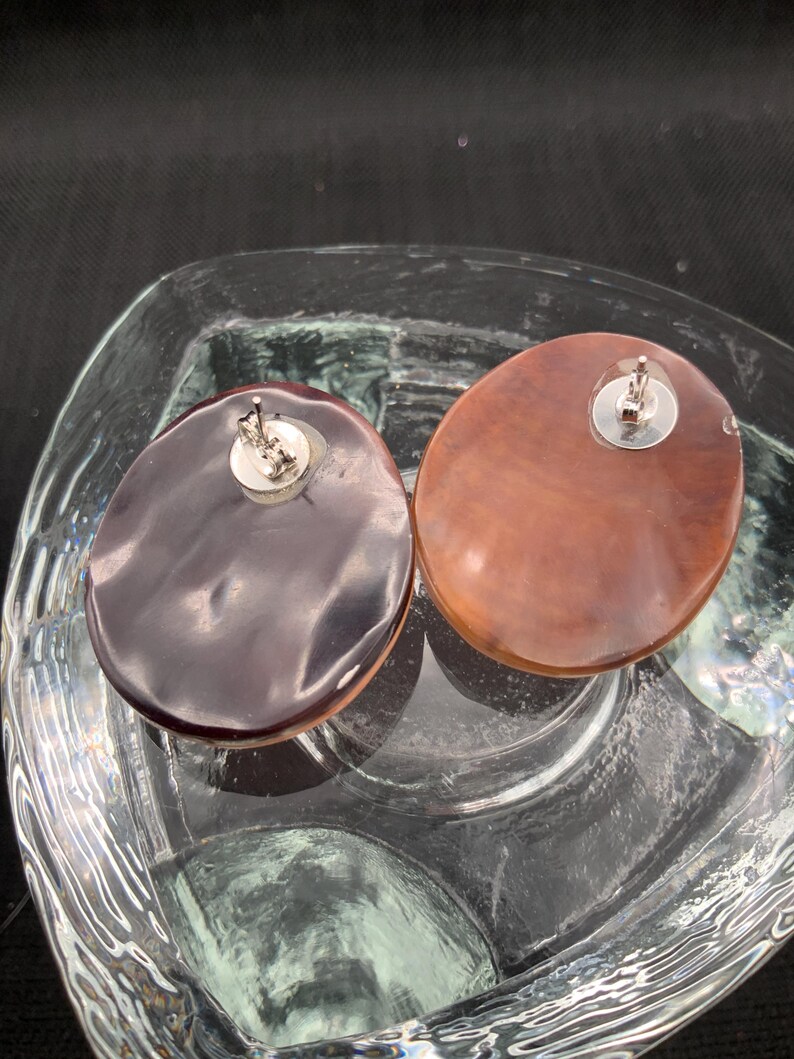 May include: Two oval-shaped stud earrings. One earring is a dark brown color, and the other is a lighter brown. Both earrings have a silver-colored post and backing. The earrings are displayed in a clear glass dish.