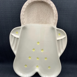 May include: A white ceramic angel-shaped dish with yellow star cutouts. The dish has a textured, beige-colored head area with a decorative swirl pattern. The wings extend outward, and the base has a double-lobed shape.