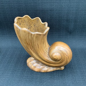 Puede incluir: Un jarrón de cerámica beige con forma de concha marina y borde ondulado. El jarrón tiene un diseño en espiral y se asienta sobre una base. El interior del jarrón tiene un patrón moteado. Es un objeto decorativo.