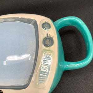 May include: A ceramic mug shaped like a retro television set. The mug features a light blue screen, a white frame, and a teal handle and accents. The mug has two small knobs and a series of vertical lines on the side.