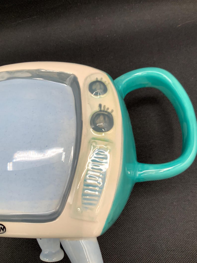 May include: A ceramic mug shaped like a retro television set. The mug features a light blue screen, a white frame, and a teal handle and accents. The mug has two small knobs and a series of vertical lines on the side.