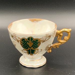 Puede incluir: Taza de té vintage con acabado blanco iridiscente, ribetes dorados y un diseño decorativo de escudo verde y dorado. El asa es dorada y ornamentada. La taza se apoya sobre una pequeña base.