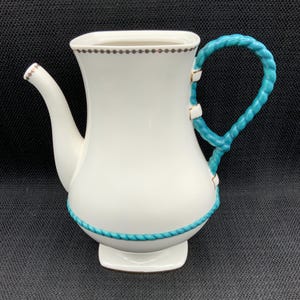 Könnte beinhalten: Weiße Keramik-Teekanne mit türkisblauem, seilartigem Griff und Zierleiste. Die Teekanne hat einen Ausguss und einen quadratischen Sockel. Der Rand ist mit kleinen braunen Punkten verziert. Ein dekoratives Stück für die Küche oder das Esszimmer.
