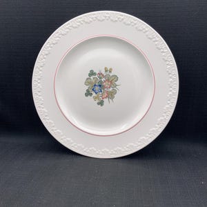 Pode incluir: Prato de jantar de cerâmica branca com uma borda elevada e um desenho floral no centro. O desenho apresenta flores rosa, amarelas, azuis e verdes com folhas verdes.