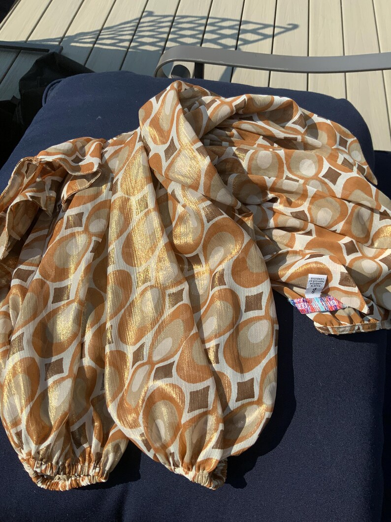 K&ouml;nnte beinhalten: Eine gemusterte Bluse in Gold- und Braunt&ouml;nen mit einem sich wiederholenden geometrischen Design aus Kreisen und Quadraten. Die langen &Auml;rmel sind an den Manschetten gerafft. Der Stoff ist ein leichtes Material in Gold-, Braun- und Cremet&ouml;nen.