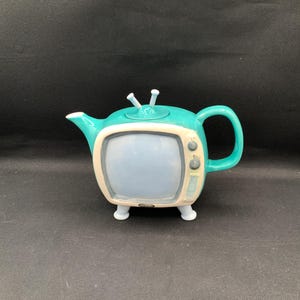 May include: A teal ceramic teapot shaped like a vintage television set. The teapot has a light blue screen, a cream-colored frame, and a handle. It features two small antennas and sits on four white feet.
