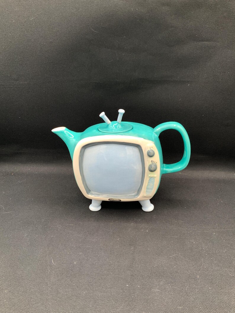 May include: A teal ceramic teapot shaped like a vintage television set. The teapot has a light blue screen, a cream-colored frame, and a handle. It features two small antennas and sits on four white feet.