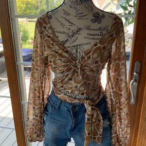 K&ouml;nnte beinhalten: Eine lang&auml;rmlige, braun-cremefarbene Bluse mit einem kreisf&ouml;rmigen Muster wird mit Jeansshorts kombiniert. Die Bluse ist an der Taille gebunden. Die Shorts sind aus blauem Denim mit ausgefransten Kanten.