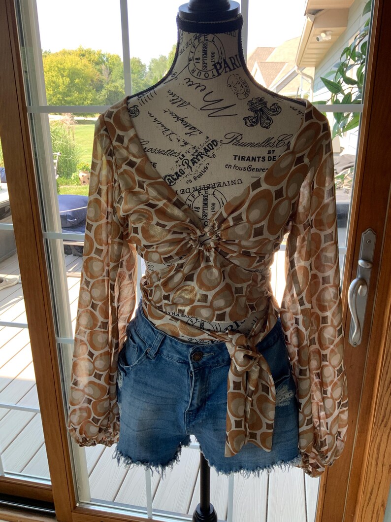 K&ouml;nnte beinhalten: Eine lang&auml;rmlige, braun-cremefarbene Bluse mit einem kreisf&ouml;rmigen Muster wird mit Jeansshorts kombiniert. Die Bluse ist an der Taille gebunden. Die Shorts sind aus blauem Denim mit ausgefransten Kanten.