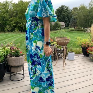 Puede incluir: Un vestido largo hasta el suelo con un estampado floral vibrante en tonos azul brillante, turquesa, blanco y verde. La prenda tiene un escote en V, mangas cortas con vuelo y un estilo cruzado que se ata al costado, creando una silueta fluida.