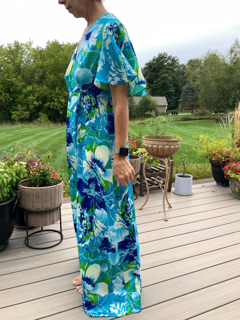 Puede incluir: Un vestido largo hasta el suelo con un estampado floral vibrante en tonos azul brillante, turquesa, blanco y verde. La prenda tiene un escote en V, mangas cortas con vuelo y un estilo cruzado que se ata al costado, creando una silueta fluida.