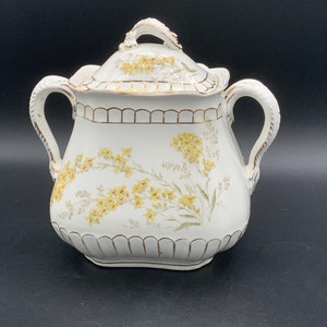 May include: White porcelain sugar bowl with a lid and two handles. The bowl is decorated with a floral pattern of yellow flowers and green leaves. The bowl has a gold trim around the top and bottom.