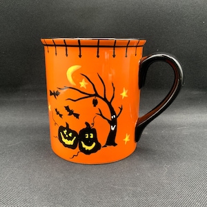 May include: Orange ceramic Halloween mug with a black handle. The mug features a spooky design with jack-o'-lanterns, bats, a tree, a moon, and stars. The rim of the mug has a black stitched design.