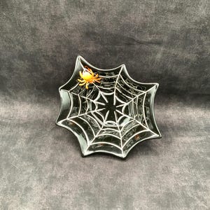 Pode incluir: Um prato preto em forma de estrela com um padrão branco de teia de aranha na superfície. Uma pequena figura de aranha laranja brilhante está fixada na parte superior esquerda do design, contrastando com o acabamento escuro e brilhante.