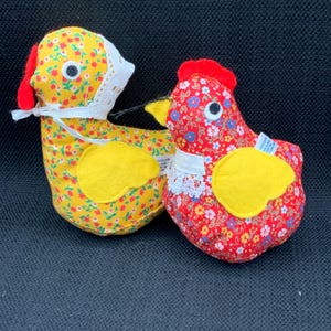 May include: Two handmade fabric chickens, one yellow with red accents and a white lace bonnet, and one red with white and blue floral accents. Both have yellow felt beaks and feet.