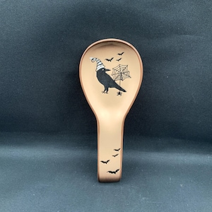 May include: A beige ceramic spoon rest featuring a black crow wearing a striped hat, holding a spider web with a small spider. The design includes several small black bats scattered across the bowl and handle of the spoon rest.