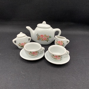 May include: A white ceramic miniature tea set with a teapot, sugar bowl, creamer, and two teacups with saucers. Each piece is decorated with a floral pattern in pink, red, and green.