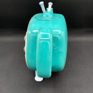 May include: A teal ceramic teapot with a rounded rectangular shape. The teapot has a handle, a spout, and a lid with two white antenna-like protrusions. The teapot is a solid teal color with white accents.