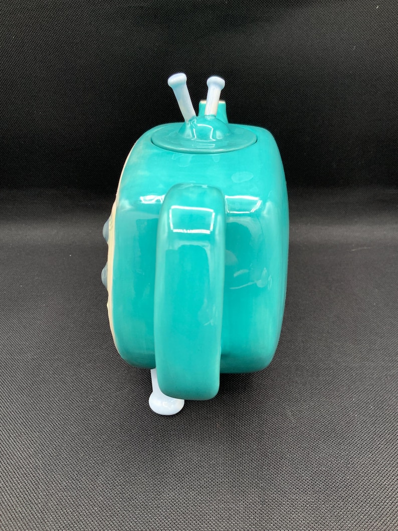 May include: A teal ceramic teapot with a rounded rectangular shape. The teapot has a handle, a spout, and a lid with two white antenna-like protrusions. The teapot is a solid teal color with white accents.