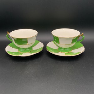 May include: Two white teacups with green and gold geometric patterns and matching saucers.