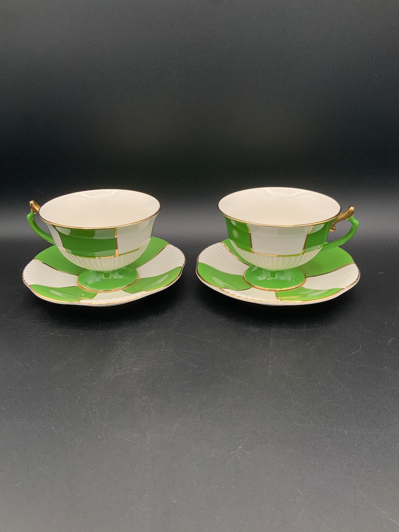 May include: Two white teacups with green and gold geometric patterns and matching saucers.