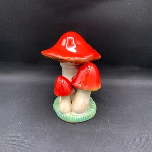 May include: A ceramic mushroom figurine with three mushrooms. The mushroom caps are glossy red, and the stems are white. The base is green with a grass-like texture. The figurine is a decorative item.