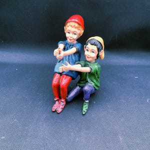 May include: A figurine of two figures sitting together. One figure wears a red hat, blue top, and red pants. The other figure wears a yellow hat, green top, and blue pants. Both figures have light skin, dark hair, and are reaching forward with arms.
