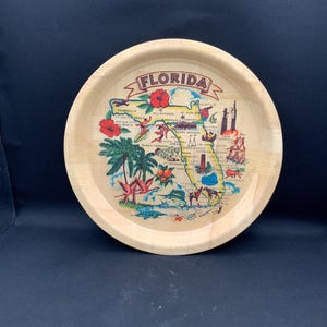 May include: A round wooden tray featuring a colourful map of Florida. The design includes the word FLORIDA in a banner, illustrations of palm trees, hibiscus flowers, oranges, a sailboat, a rocket, and various wildlife like a lion and a fish.