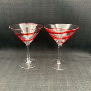 Puede incluir: Dos copas de martini transparentes con rayas rojas. Las copas tienen una forma de cuenco cónico clásico y un tallo largo. El borde de cada copa también está delineado en rojo. Fondo oscuro.