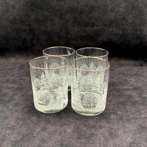May include: A set of four clear glass tumblers featuring a frosted white etched design of pine trees and falling snow. Each glass has a thin gold-colored rim around the top edge, providing a decorative accent to the winter-themed glassware.