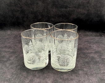 Könnte beinhalten: Ein Set aus vier klaren Trinkgläsern mit einem mattierten, weißen Ätzdesign aus Tannenbäumen und fallendem Schnee. Jedes Glas hat einen dünnen, goldfarbenen Rand an der Oberkante, der einen dekorativen Akzent für die winterlichen Gläser setzt.