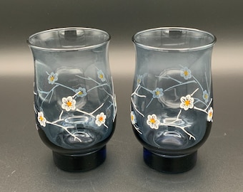 Könnte beinhalten: Ein Paar Trinkgläser aus durchscheinendem rauchgrauem Glas mit einem aufgemalten Motiv aus weißen und hellblauen Kirschblüten auf dünnen, weißen Zweigen. Jedes Glas hat einen geschwungenen, ausgestellten Körper und einen massiven, dunkelblauen Boden.
