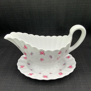 May include: White porcelain gravy boat with a matching saucer. The boat has a scalloped rim and a handle. Both pieces are decorated with small pink and blue floral patterns. A classic addition to any table setting.
