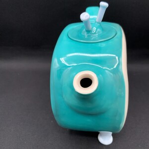 May include: A teal ceramic teapot with a rounded body and a white accent on the side. The teapot has a spout, a lid with two white knobs, and a small white foot. The teapot is a unique and decorative kitchen item.