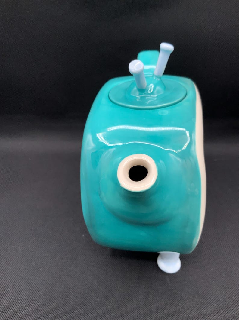 May include: A teal ceramic teapot with a rounded body and a white accent on the side. The teapot has a spout, a lid with two white knobs, and a small white foot. The teapot is a unique and decorative kitchen item.