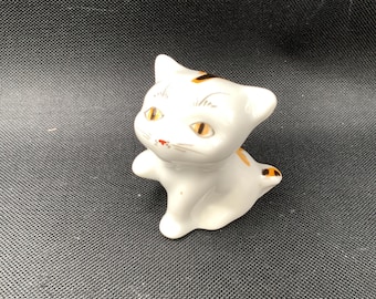 May include: A small white ceramic cat figurine featuring painted orange and black markings on its head and tail. The cat has yellow eyes, a small red nose, and delicate whiskers painted on its face, with one front paw slightly raised.